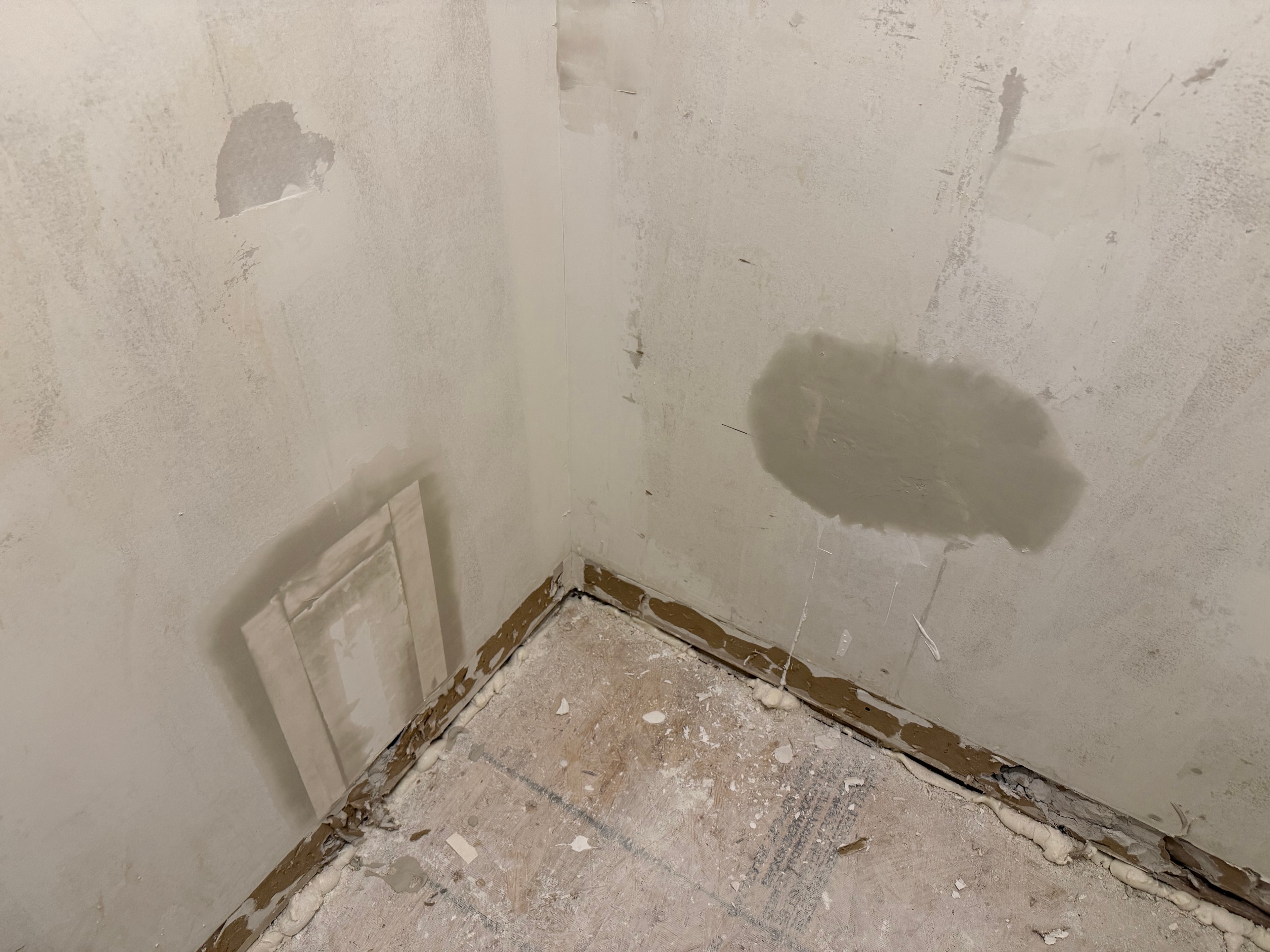 Bathroom corner showing patched walls and bare subfloor mid-renovation
